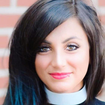 Photo of Elle a white woman with long dark brown hair with a lip ring on the left side of her lower lip and fuschia lippy. She is wearing a clerical collar and smiling slighty into the camera.