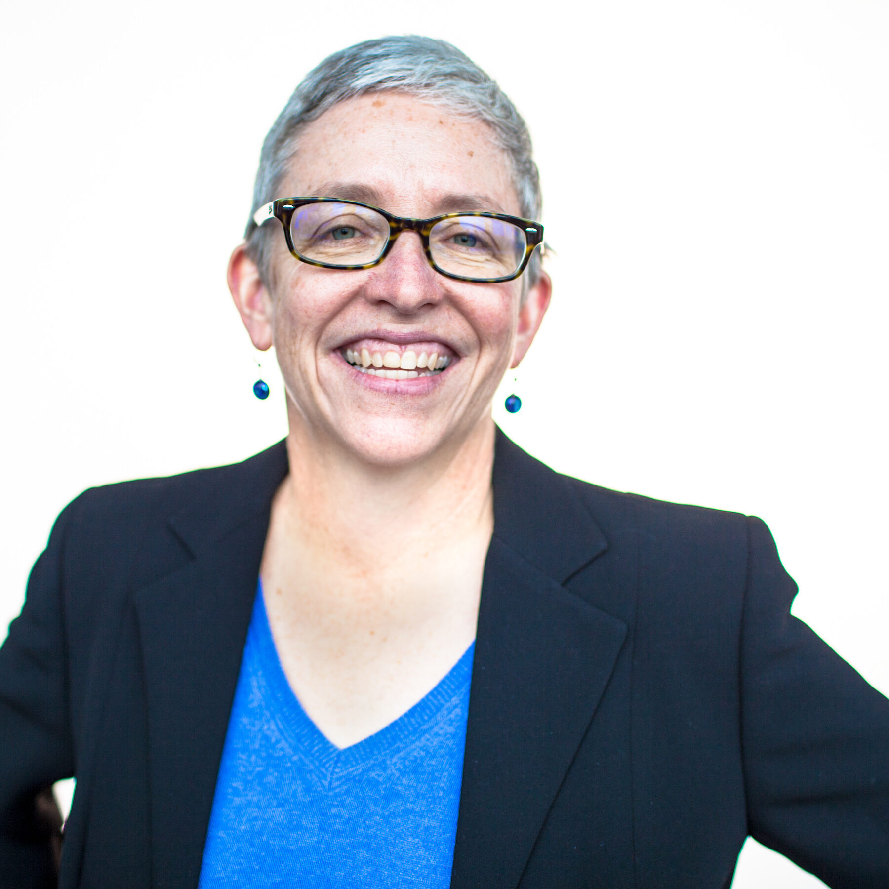 Photograph of Shari, a white woman with short silver hair wearing dark rectangular glasses. She is smiling and wearing a dark blazer over a blue v-neck shirt.