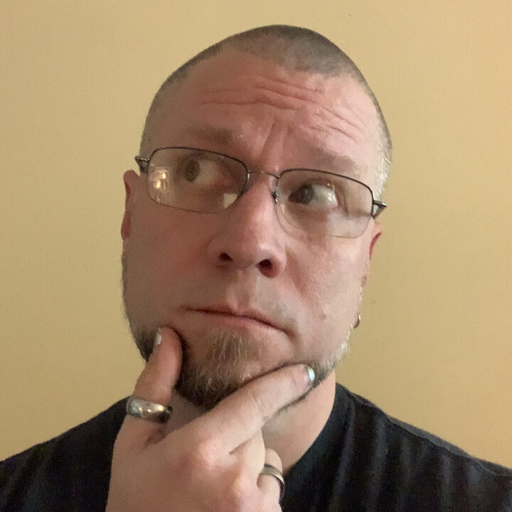 Photo of Jaye, white human with a buzz cut, short brown goatee and wire rimmed glasses. They are holding their thumb and forefinger to thier chin making a quizzical expression. They are also wearing a thumb ring and silver nailpolish.