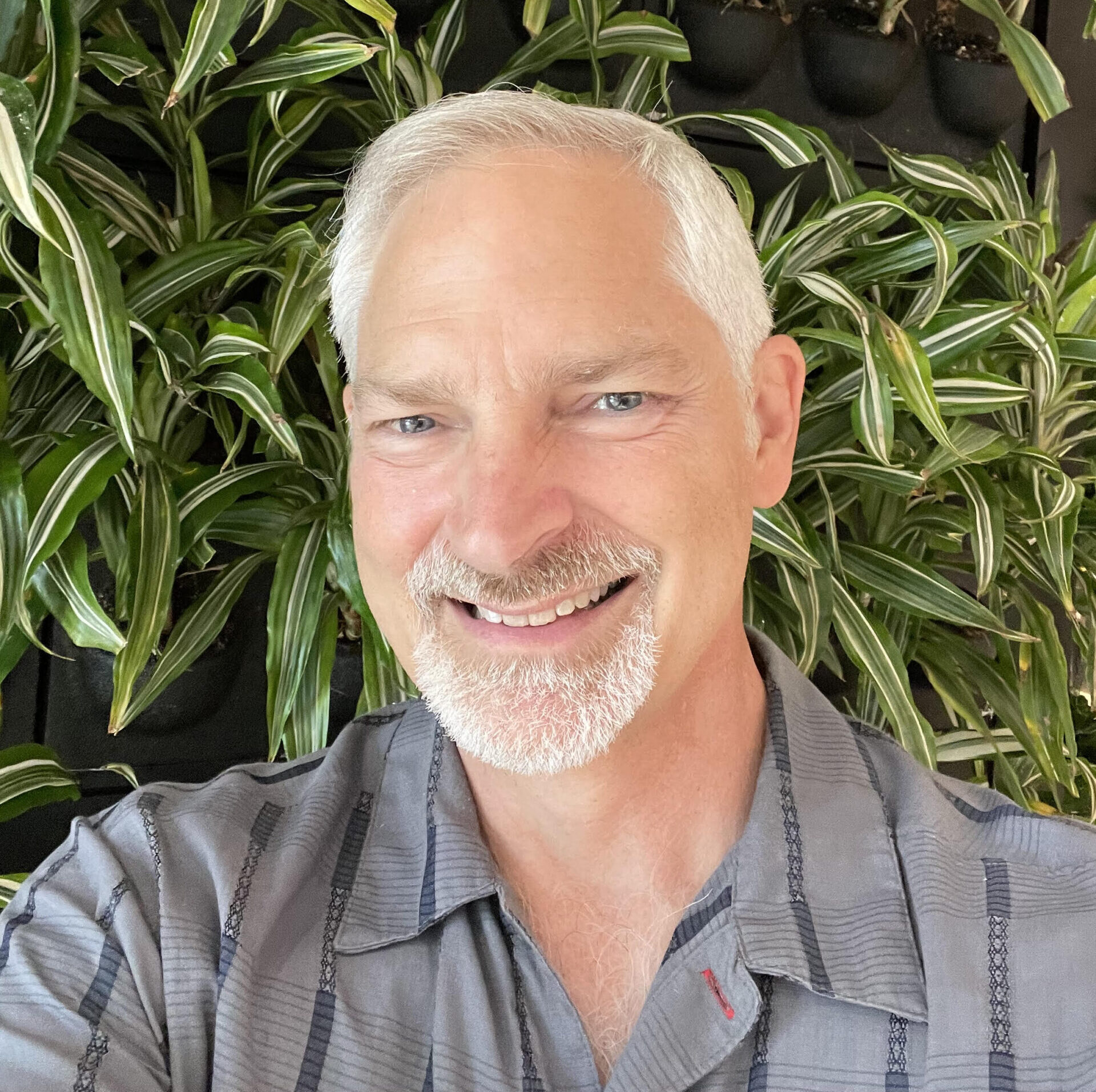 Headshot of Michael Wilker. Michael has short white hair with a bald patch on top, white/grey goatee, blue eyes, and peach white skin. He is smiling, and wearing a grey button down with grey stripes and standing in front of green plants.